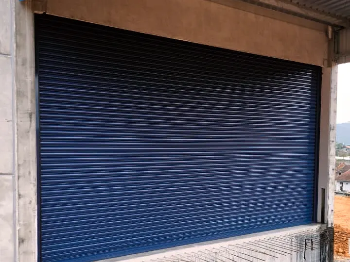 Porta de enrolar azul escura instalada no vão de um galpão em construção, sobre mureta de concreto com ferragem aparente