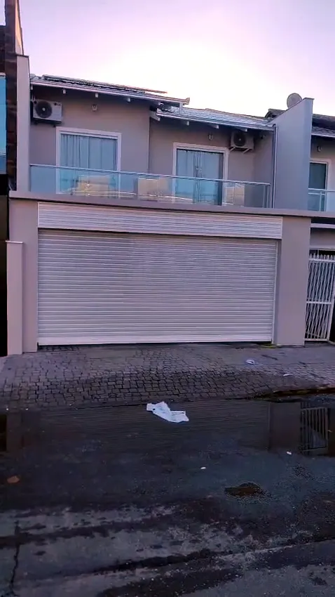 Porta de enrolar clara subindo na garagem de um sobrado, filmada da calçada no fim da tarde
