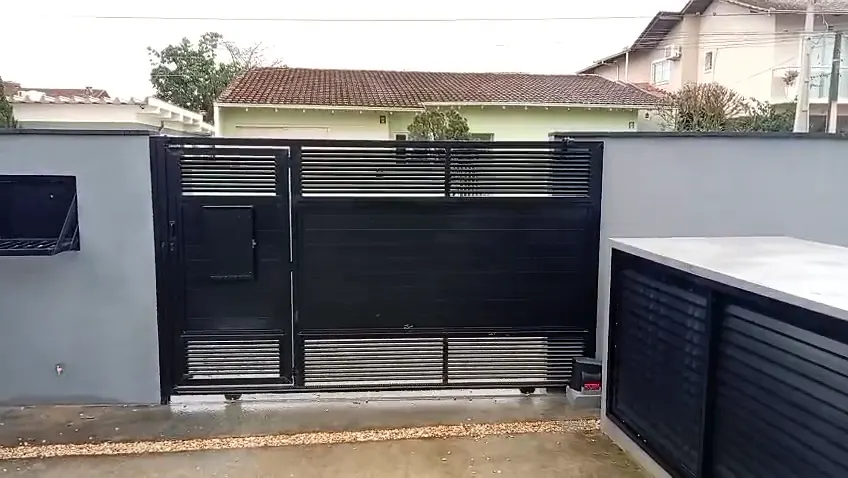 Portão de correr preto ripado abrindo, com porta social embutida na folha e abrigo de lixeira em ripado ao lado