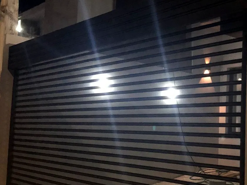 Portão ripado horizontal escuro instalado na fachada de uma casa, com a luz da garagem acesa atrás das ripas