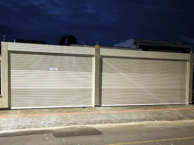 Duas portas de enrolar bege instaladas lado a lado na fachada de um ponto comercial, fotografadas à noite